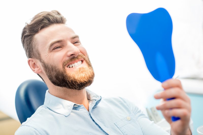 a male patient with a beard and smiling in the handheld mirror while at the dentist’s office