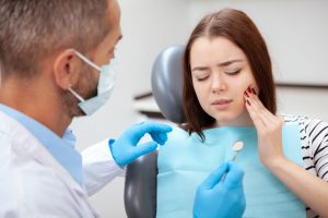 Woman with brown hair sitting in dentist's chair holding her hand to her cheek in pain